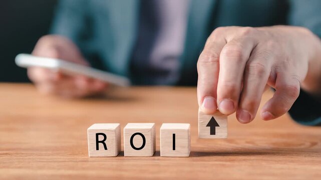 Close-up of a businessman stacking wooden blocks to reveal ROI text and a rising arrow icon, holding a smartphone for financial investment return, business growth, and profit analysis concept.