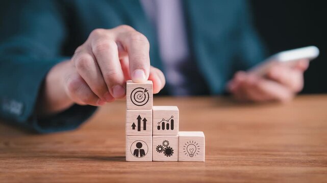 Close-up of a businessman stacking wooden blocks with innovation, workflow, and target icons, holding a smartphone for corporate strategy planning, business growth, and organization success concept.