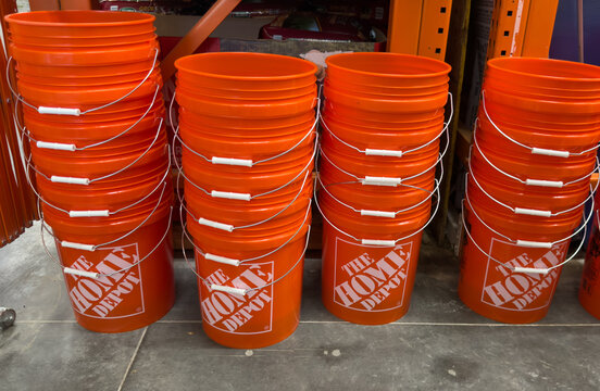 Stacks of iconic bright orange Home Depot buckets arranged neatly in rows on a concrete floor inside a bustling retail hardware and home improvement store, ready for purchase.