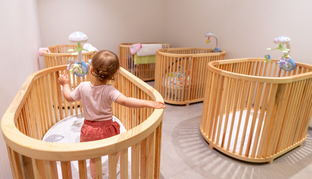 Toddler standing and playing in crib in modern nursery filled with some wooden cribs