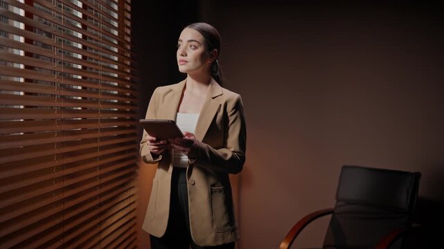 Woman standing near window blinds. Female working with tablet at dim room. Woman wearing professional clothing and holding tablet. Loft style of dark room. Film agency coworker using device.