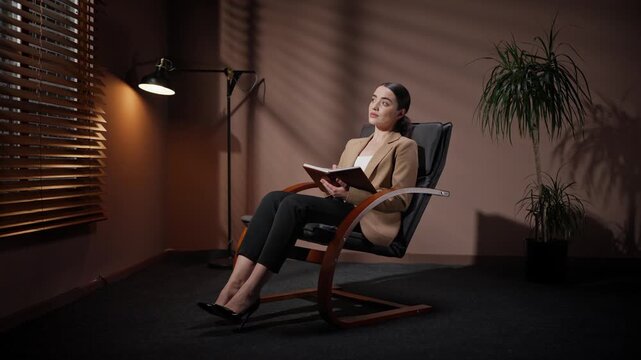 Thoughtfull professional woman sitting on chair. Female in official outlook isolated in dark office room. Woman occupied by office lamp and flowerpot. Female making business notes late in evening.