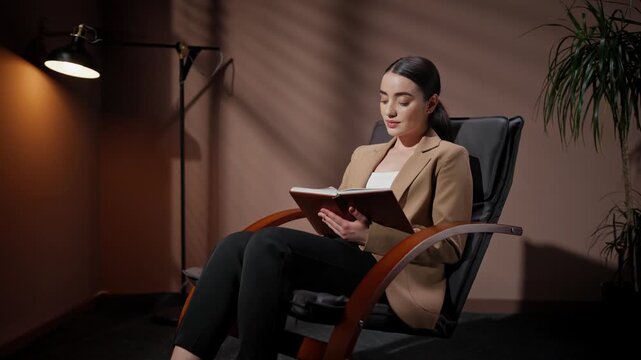 Thoughtfull professional woman sitting on chair. Female in official outlook holding notebook. Woman occupied by office lamp and flowerpot. Female making business notes indoor.