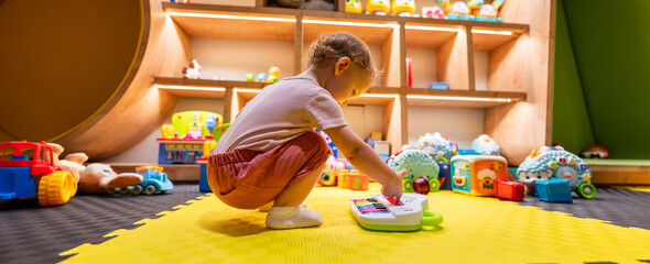 Toddler playing with colorful musical toy piano in a bright playroom filled with educational toys and activities for early childhood development © Maria