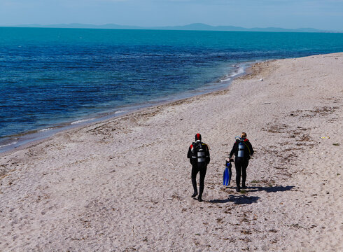 Two divers in wetsuits walking along a sandy beach carrying diving gear, with clear turquoise water and distant hills visible in the background