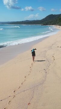 Walking along vibrant coastline. Serene woman strolls by emerald water with gentle waves. Peaceful woman walks along turquoise shore with reflected sky and distant forest, vertical video