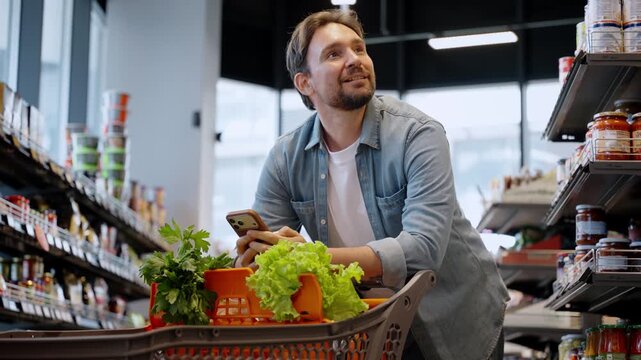 Happy bearded man texts on smartphone as leans on shopping cart