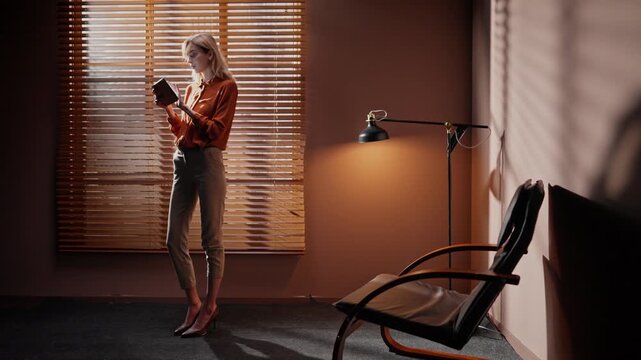 Woman isolated at background of window blinds. Female standing occupied by office lamp and chair. Woman wearing professional clothing and holding tablet. Loft style of dark room.