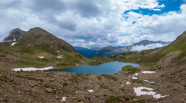 Panoramic view of a Tristaina glacial lake in Andorra, teal mountain lake nestled in a rocky alpine cirque with snow patches, rugged Pyrenean peaks and dramatic clouds above a deep valley