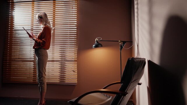 Zoom shot of woman standing near window with blinds. Woman isolated at office room. Casting director at background of office furniture. Woman shadow falling on wall. Room in loft style and woman.