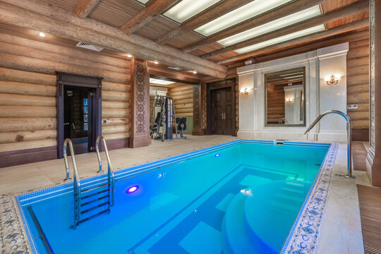 A pool is at the center of a room with wooden walls. Bright lights are on the ceiling. A fitness area is on one side of the room. There are also stairs leading into the water
