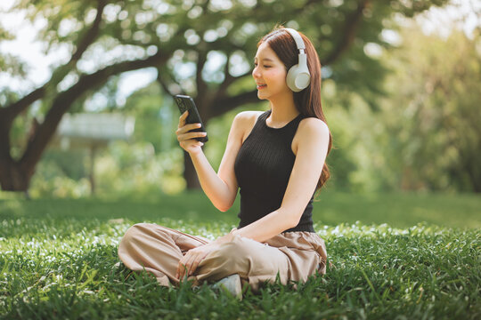 Happy Asian woman wearing headphones and using smartphone while sitting on green grass, smiling female listening to music, podcast, or streaming online video in public park. wellness, and technology