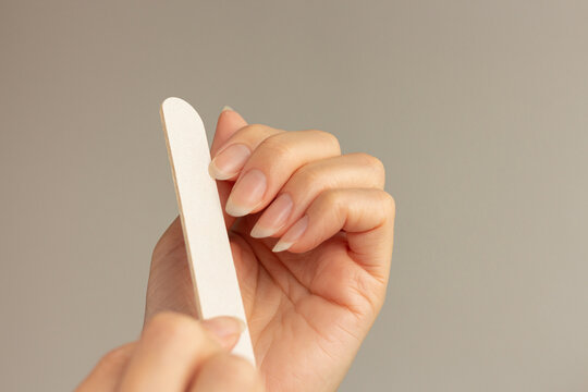 Close-up of a young woman filing her natural fingernails with a nail file, showing the process of shaping natural long nails against a minimalist neutral beige background.