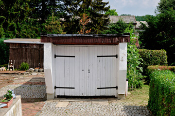 Naklejka premium White wooden double garage doors with black hinges in a green backyard garden setting