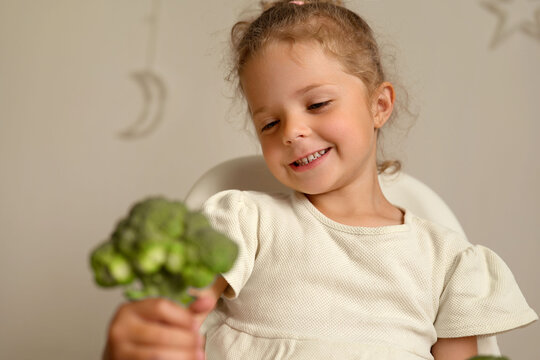 A young child with light brown hair sits in a chair and holds broccoli with a smile. The background is simple and bright, showing a pleasant home setting