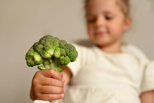 A child smiles while holding a large piece of broccoli in their hand. The setting appears to be a kitchen with a neutral background