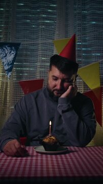 A sad, lonely man in a party hat celebrates his birthday alone, looking at a cupcake with a candle. He listlessly blows a party horn. Vertical video.