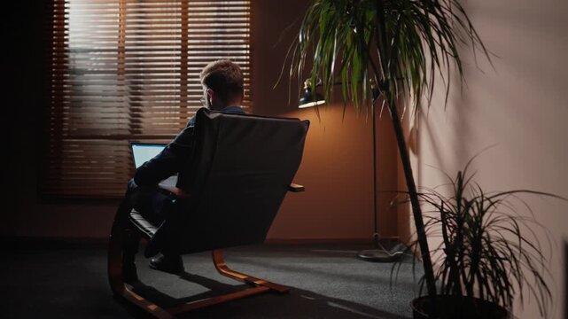 Back view of businessman sitting on armchair. Back shot of male boss sitting in front of window with blinds. Company director working late in evening. Man using laptop in dim loft room, flowerpot.