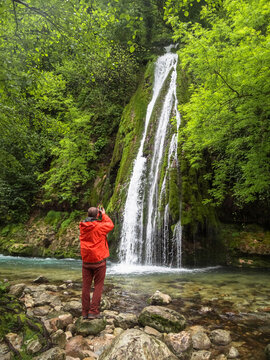 Tourist in orange jacket taking photo of Kaghu waterfall in Georgia.