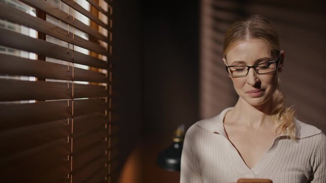 Woman in glasses preparing for audition. Woman in dark room. Female repeating script before casting. Woman holding phone and reading role. Woman standing near windows with blinds.