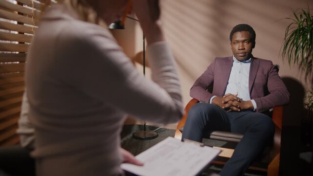 African American man auditioning for movie role. Man isolated in dark room with blinds. Female director conducting casting. Male passing film audition. Casting director holding folder.