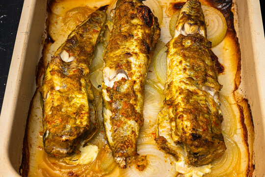 Baked fish fillets seasoned with spices and herbs, placed on a bed of sliced onions inside a rectangular ceramic baking dish