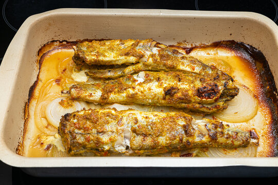 Baked fish fillets seasoned with spices and herbs in a ceramic dish, garnished with onions, resting on a stovetop, ready for serving