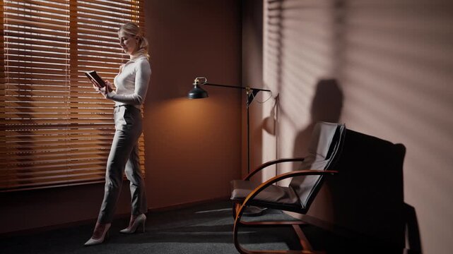 Woman standing in dim sunny room. Female wearing heels holding tablet. Casting director standing in audition room. Woman at background of window blinds and office furniture.