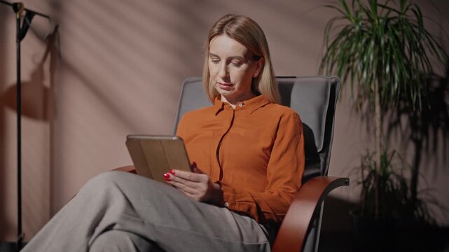 Woman working late in evening. Female sitting on chair in dim room. Woman working with tablet. Female isolated in dark loft room. Woman in bright orange shirt. Female near lamp and flowerpot.