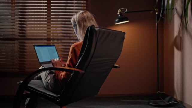 Back view of woman working with laptop. Back shot of woman sitting on chair. Female using pressing keyboard buttons. Back vision of woman workink late in evening. Dim modern equipped room.