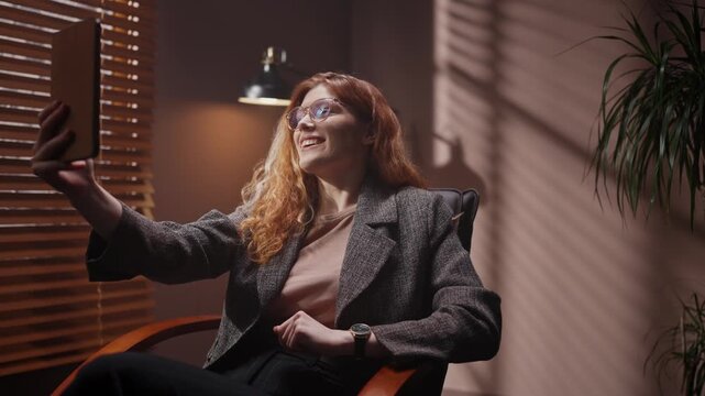 Red head woman cheerfully communicating by zoom chat. Female holding tablet and having video call. Happy female smiling with teeth in laptop screen. Woman gesturing sitting on chair in dark room.
