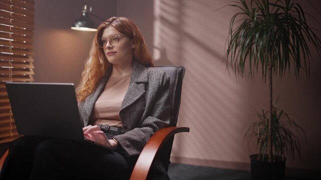Red head woman looking at tablet screen. Female isolated in dark room. Woman wearing jacket and smiling. Female sitting on chair isolated in dim room. Woman in glasses satisfied with working.