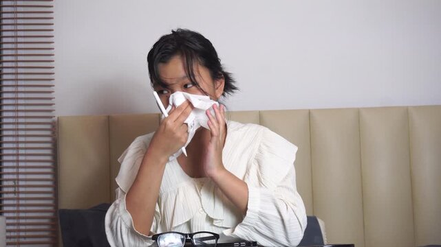 Teenage girl using tablet on bed at night while sick. Asian student blowing nose with tissue paper inside bedroom during her ill Gen Z lifestyle remote job winter flu cold disease.