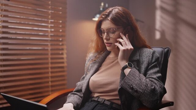 Businesslady having business call. Woman wearing glasses sitting on chair. Red head female using laptop keyboard. Woman communicating by smartphone. Female working with two gadgets.