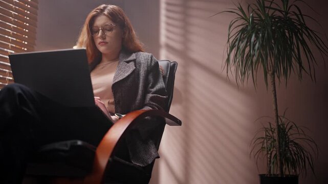 Red head woman looking at laptop screen. Female isolated in loft room. Satisfied woman wearing jacket and glasses. Female sitting on chair clicking laptop keyboard. Woman working late in evening.