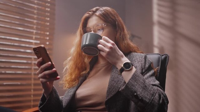 Red head woman wearing official jacket and glasses. Female relaxing during work break. Woman holding smartphone and scrolling social media ribbon. Businesslady drinking cup of tea or coffee.