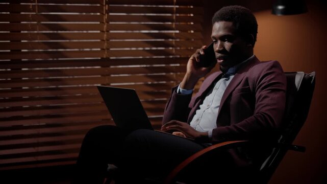Business man sitting on chair in dim room. African American man conducting business call. Male working with laptop in dark room. Male working with devices late in evening. Business man wearing suit.