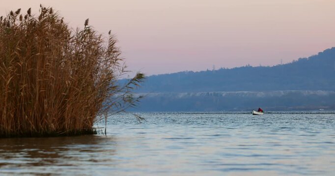 Fisherman In Small Fishing Boat On Calm Lake Near Golden Reeds At Sunset With Distant Hills and flying flock of water birds small ducks