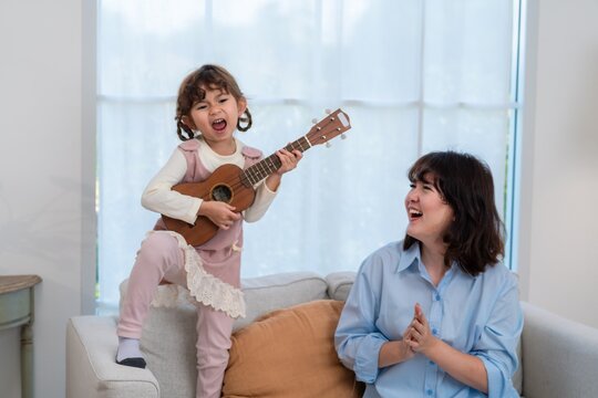 Child plays ukulele while mother smiles in living room during daytime