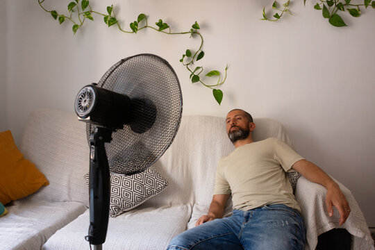 Exhausted Hispanic man resting on sofa with fan during summer heatwave