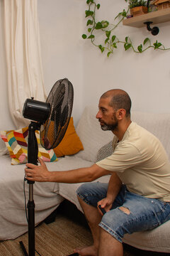 Exhausted Hispanic man reaching to switch on electric fan during heatwave