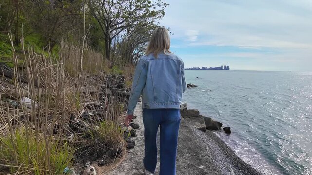 Woman wearing a denim jacket and jeans walking along a concrete embankment by the sea, with a modern city skyline visible in the distant background on a sunny day