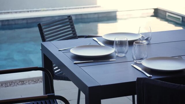 Rectangular dark patio table set for two beside pool, with two white ceramic plates, wine glasses, clear water glass, cutlery, folded napkin, and textured surface in bright daylight, creating calm