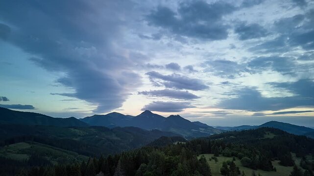 Panoramic mountain dusk landscape with jagged central peak silhouetted against glowing pastel sky, layered cloudbank, forested hills, rolling meadows, and distant ridgelines