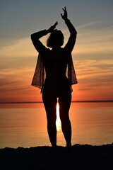 Fototapeta premium Female silhouette with hands raised in celebration on the shore at sunset, summer vacation concept.