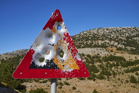 Shot triangular warning sign, large bullet holes, mountains, blue cloudless sky, Sfakia, Chania province, Crete, Greece