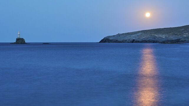 Full moon, moon over rocky coast, ray of moonlight on sea, sea calm, sky blue and cloudless, lighthouse on tiny rocky island shines, Andros Island, Cyclades, Greece