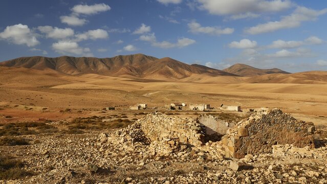 Island interior, barren landscape, reddish brown hills, ruins of buildings further away, ruin in foreground, blue sky, white clouds, Fuerteventura, Canary Islands, Spain