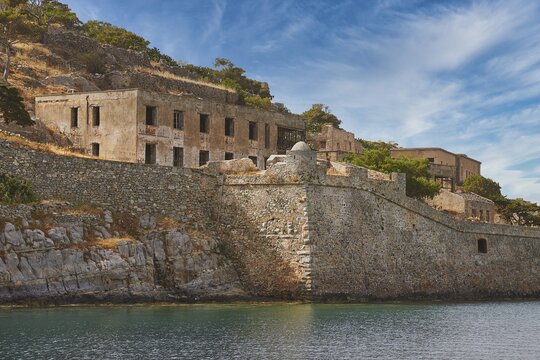 Dilapidated-looking old buildings on a rocky coastline surrounded by clear skies and calm seas, Venetian Sea Fortress, Leper Island, Spinalonga, Elounda Bay, Mirabello Bay, East Crete, Crete, Greek Islands, Greece