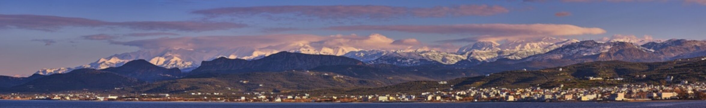 Panoramic shot, town at the foot of snow-capped mountains glowing in the sunset light, with dramatic clouds in the sky, Lefka Ori, White Mountains, mountain massif, west, Gulf of Kissamos, Crete, Greece